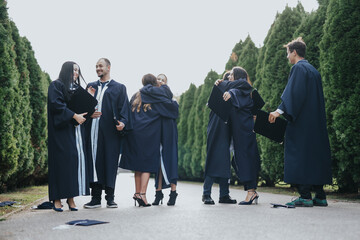 University students, in graduation gowns and caps, celebrate success in a sunny park. They walk, smile, congratulate each other, and throw caps in the air, creating positive memories.