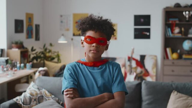 Portrait of confident African American boy wearing superhero cape and mask turning to camera and posing with arms crossed in living room at home