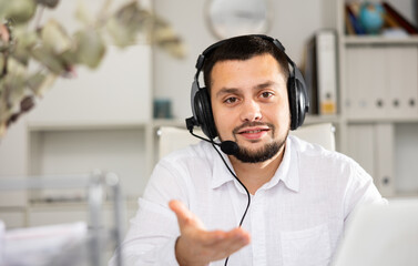 Portrait of positive call center worker wearing headphones with microphone, explaining with gesture, looking at camera.