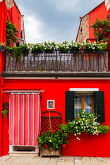 Bright traditional red house on Burano island, Venice, Italy. Colorful curtain on door, wooden old style window with shutters and Mandevilla flowers on window sill.