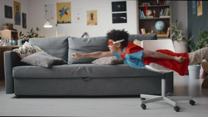 Slow motion shot of African American father pushing little son in superhero costume on chair through living room, kid holding one arm straight out and smiling while imitating flight