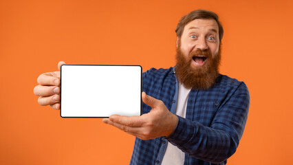 Excited redhaired bearded guy showing digital tablet empty screen, studio