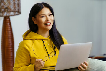 A woman in a yellow hoodie is smiling and holding a laptop. She is wearing glasses and she is enjoying her time