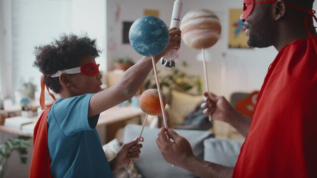 Little Black boy and his father dressed in superhero costumes playing with toy rocket and planets at home