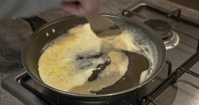 Scrambled eggs being cooked and stirred in a frying pan on stove hob with a wooden kitchen utensil. Culinary food preparation, breakfast meal