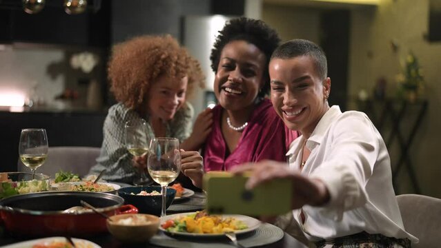 Three diverse young women taking selfie at home dinner table