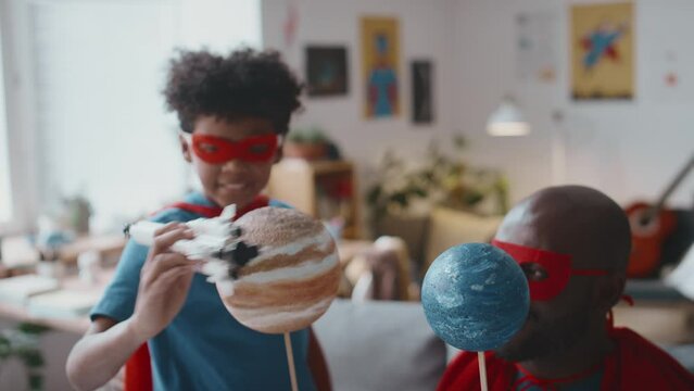 Joyous African American boy in superhero cape and mask playing with toy rocket imitating its flight among planets held by father while having fun at home