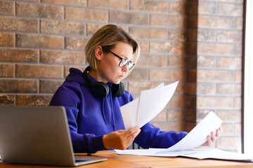A young woman in a blue sweater is rearranging papers on a table, an open laptop. Girl in glasses working in office with papers, bank employee, student doing assignments, girl studying