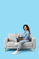 Obraz premium Young African-American woman using laptop on sofa against blue background