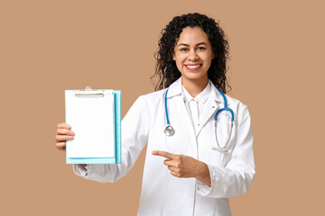 Beautiful young happy African-American female doctor with stethoscope pointing at clipboard on brown background