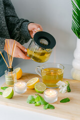 A hand pouring herbal tea surrounded by refreshing citrus, a glowing candle, and lush greenery, creating an inviting atmosphere for relaxation.