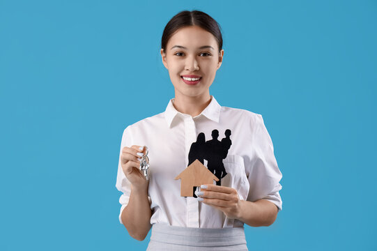 Asian real estate agent with figures and keys from house on blue background - Powered by Adobe