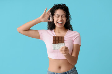 Beautiful young African-American woman in headphones with sweet chocolate bar on blue background - obrazy, fototapety, plakaty