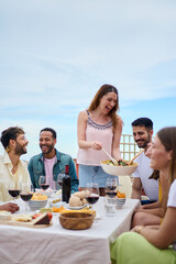 Vertical. Group of people gathered around table, sharing food and wine while smiling and enjoying leisurely event. The hostess serves food to her guests. Friends lunch together on a spring summer day