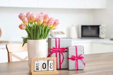 Calendar with date of International Women's Day, gift boxes and tulips on table in kitchen