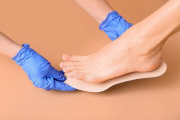 Doctor hands in medical gloves fitting orthopedic insoles to female legs on beige background, closeup