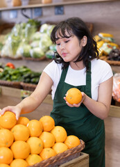 Focused interested young asian salesgirl in green apron arranging fruits on produce display in...
