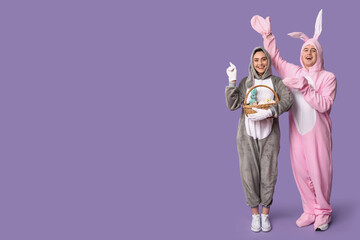 Beautiful young couple in bunny costume with Easter basket pointing at something on purple background