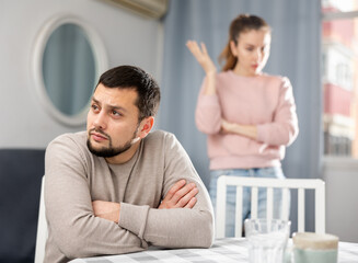 Upset bearded guy sitting at table in living room while frustrated girlfriend scolding him in...