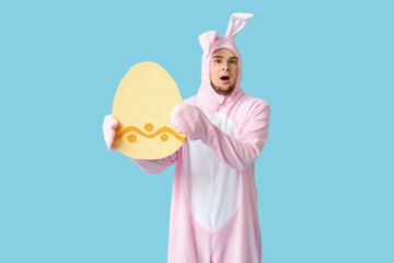 Shocked young man in bunny costume with paper Easter egg on blue background