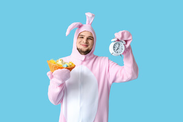 Young man in bunny costume holding wicker basket with Easter eggs and alarm clock on blue background