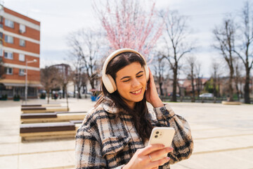 One young girl is listening to music on her wireless headphones and using her phone outdoors on a sunny day
