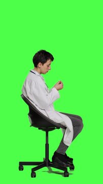 Profile Physician in white coat waiting for patients at consultations, feeling impatient sitting on a chair against greenscreen backdrop. Medic practitioner with stethoscope waits for people. Camera A