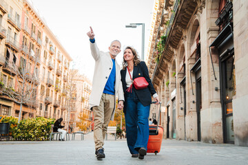 Mature tourists pointing and walking with a suitcase on vacations. Middle aged couple having fun...