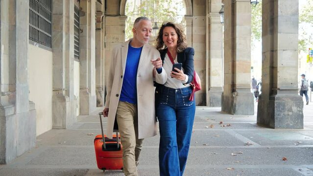 Mature tourists pointing and walking with a suitcase on vacations. Middle aged couple having fun sightseeing on a european downtown. Real mid adult married people enjoying a journey trip on holidays.