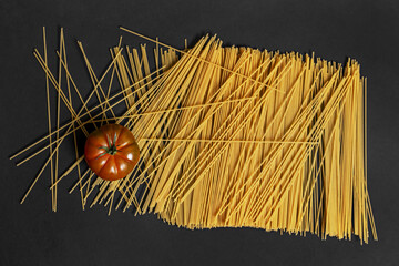 Bright raf tomato on a large pile of raw wheat spaghetti spread on a smooth black surface - obrazy, fototapety, plakaty