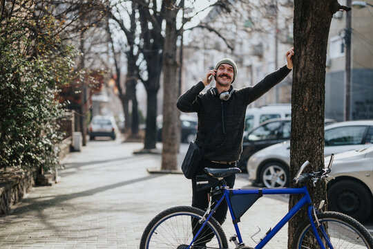 Urban lifestyle scene with a joyful man chatting on a smart phone and leaning against a tree beside his bike on a bright day.
