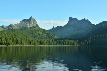Reflection of high pointed rocks with gentle banks overgrown with dense taiga in the waters of a picturesque mountain lake on a sunny summer day.