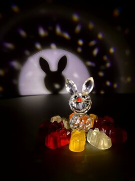 Theatrical pictures of a small doll made of glass surrounded by gummy bears with lighting and a black background (not made by ai)