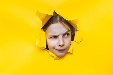 A funny red-haired girl looks out through a torn hole in yellow paper. The concept of surprise, fear, fright, and skepticism from what he saw. Discounts, sales, surprise. Copy space.