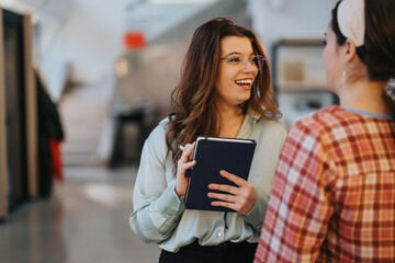 Two female colleagues are having a jovial discussion in a business setting, one holding a digital...