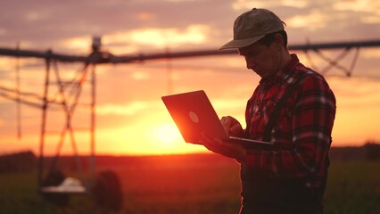 silhouette agriculture. a male farmer works on a laptop in a field with green corn sprouts. corn is watered by irrigation machine. sunlight irrigation agriculture business concept - 767536616