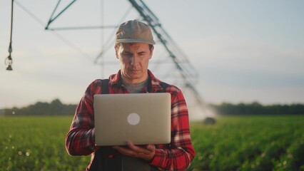 corn agriculture. a male farmer works on a laptop in a field with green corn sprouts. sunlight corn is watered by irrigation machine. irrigation agriculture business concept - 767536648