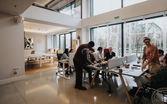 Inclusive modern office setting with a diverse team engaging in discussion. Wheelchair user and coworkers interact around computers.
