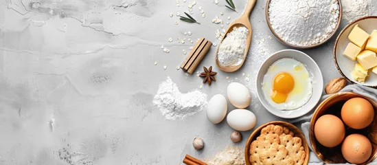 Fototapeten Baking ingredients like flour, eggs, butter, and kitchen textiles laid out on a bright grey concrete background for a cookies, pie, or cake recipe mockup with copy space in top view. © Vusal
