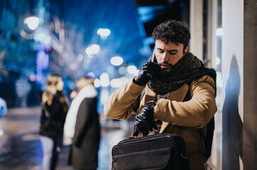 A pensive young adult male is seen interacting with his smart phone on a bustling city street at...