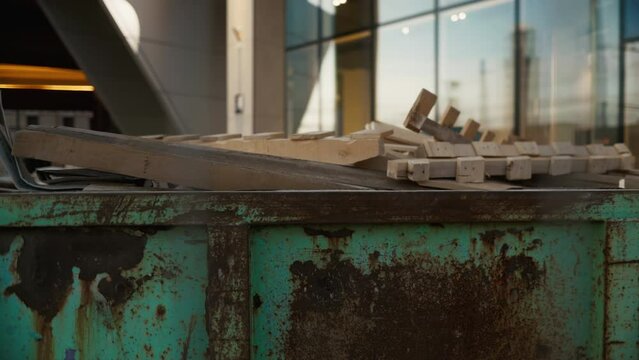 A green dumpster full of wooden pallets by a building