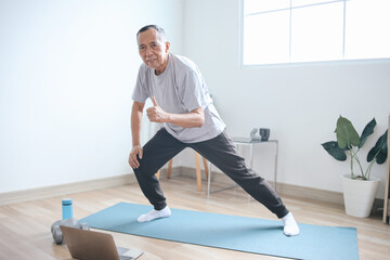 Obraz premium Senior Asian man exercising using laptop, having online fitness class from home and showing thumbs up
