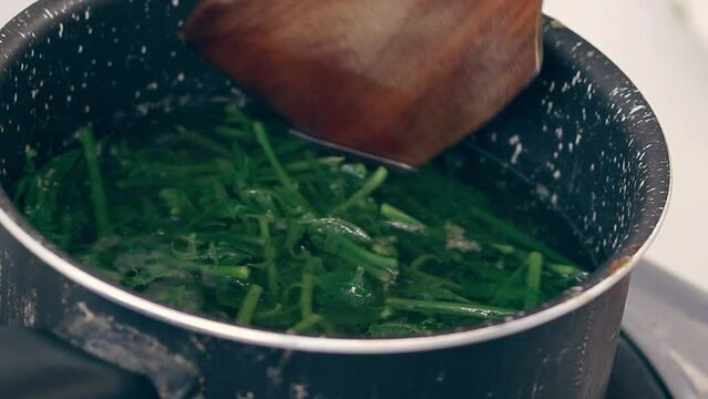 Using a wooden spatula to stir pot of boiling water blanching fiddlehead ferns for making pako salad, a local Filipino delicacy showing the authentic daily home life and culinary heritage