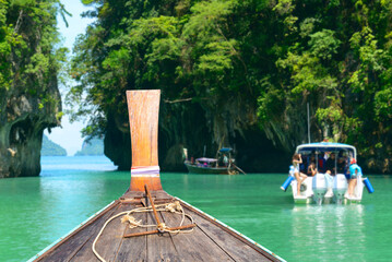 Wooden longtail boat prow on emerald waters with tourists and limestone cliffs in the background..