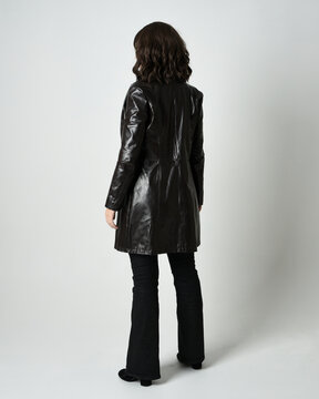 Full length portrait of brunette woman wearing long leather trench coat and black boots. Standing pose walking away from camera, facing backwards. Isolated silhouette on white studio background.