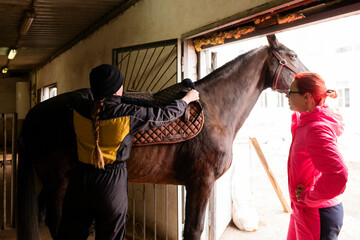 Two people saddling a brown horse in stable.