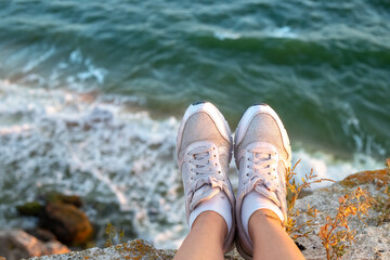Travel and tourism. Female legs in white sneakers hanging from a rocky shore on the sea coast