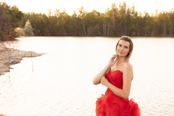 Amidst the soft hues of a tranquil lakeside at dusk, a woman in a bold red dress exudes a serene...