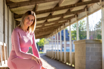 An athletic woman in pink sportswear finds a moment of quiet rest on a wooden bench under the...