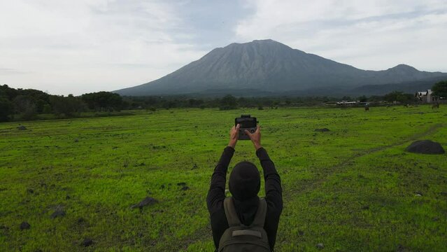 A man traveler in a hat, with a backpack, takes a photo of a mountain on his phone, rear view, Bali island, Agung volcano.